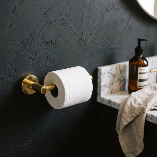 Unlacquered brass toilet paper holder with a white roll mounted on a dark textured bathroom wall beside a vanity