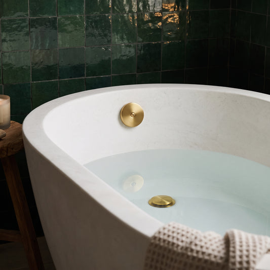 Unlacquered brass drain stopper and overflow plate inside a water-filled white bathtub against dark green tiles