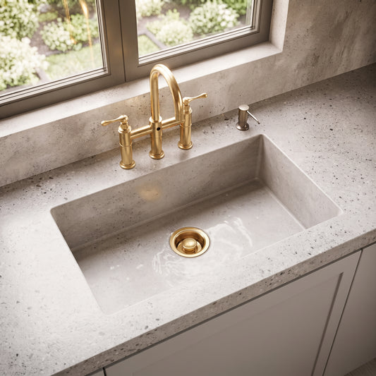 Unlacquered brass garbage disposal flange and stopper installed in a kitchen sink beneath a window and brass bridge faucet