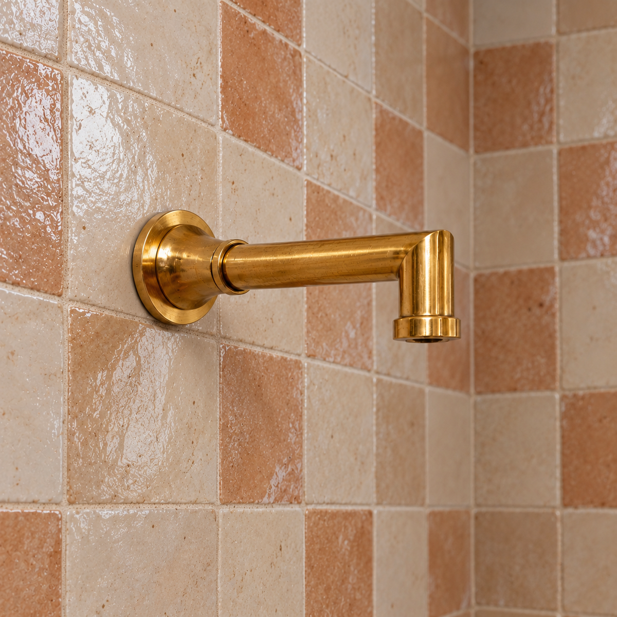 A polished brass wall-mounted shower arm with a round backplate, shown installed against a checkered beige and terracotta tile wall.
