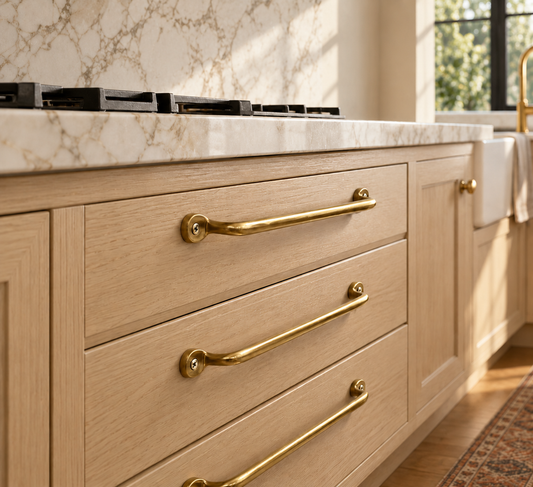 Unlacquered brass drawer pulls mounted horizontally on light wood kitchen cabinetry beneath a marble countertop