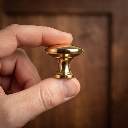A hand holding a polished brass cabinet knob against a dark brown wooden background.
