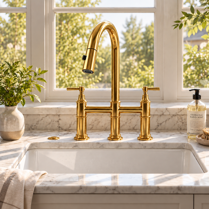 Front view of an unlacquered brass bridge kitchen faucet above a white sink beneath a sunlit window
