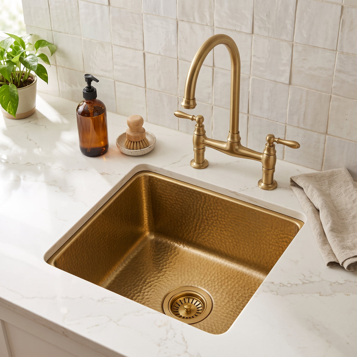 Hammered unlacquered brass prep sink undermounted in a white countertop beneath a brass bridge faucet and tiled backsplash