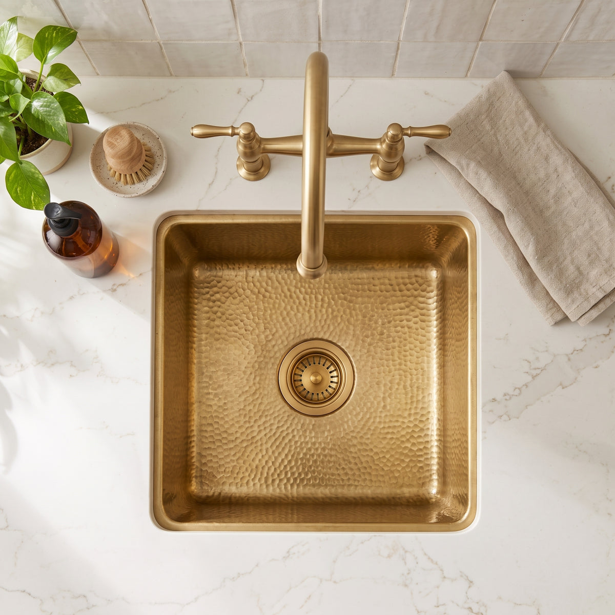 Overhead view of the hammered brass bar sink and matching strainer drain in a white countertop with a bridge faucet