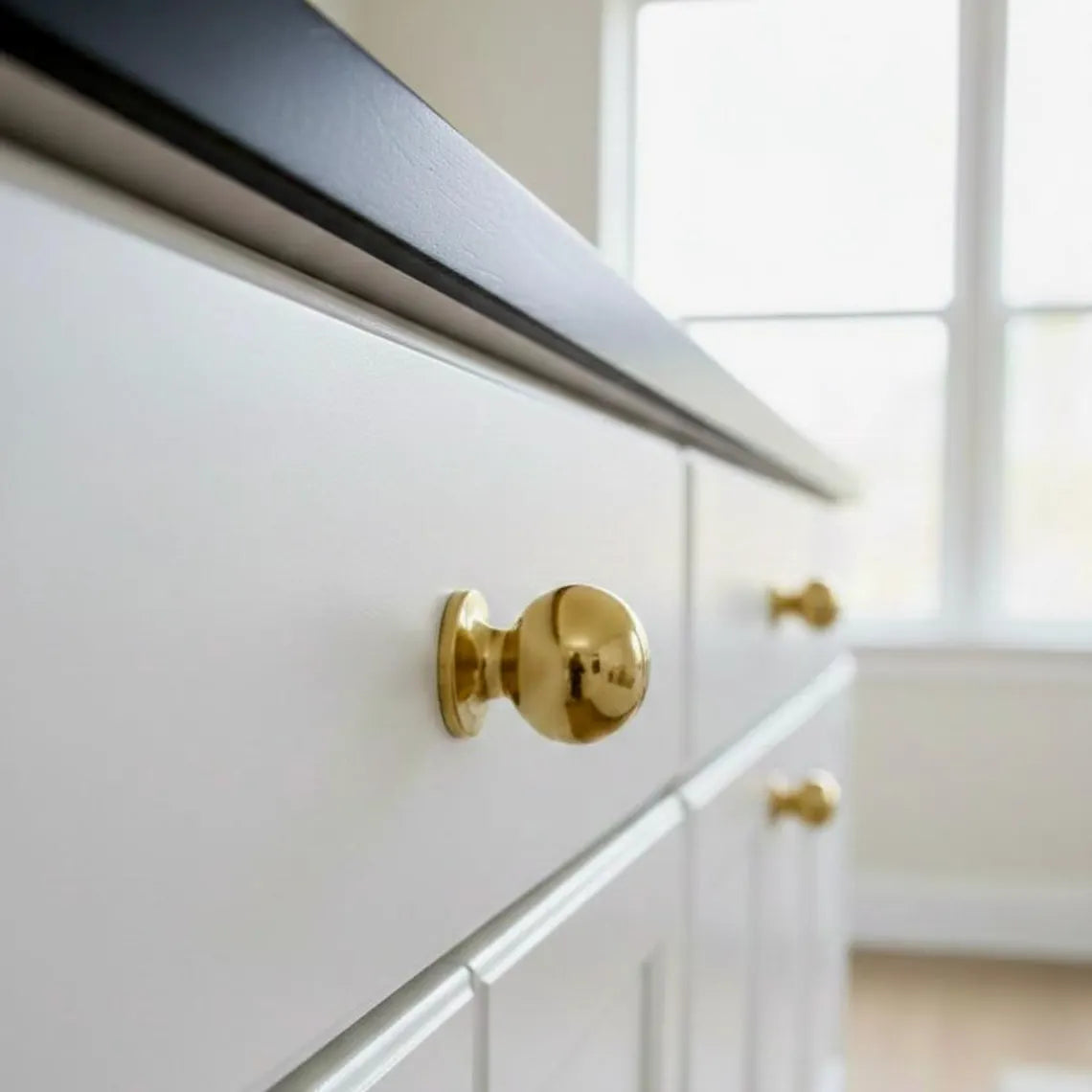 Side close-up of an unlacquered brass ball cabinet knob on a white drawer, showing its rounded head and stepped base