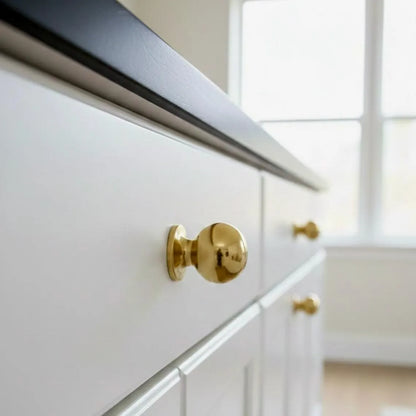 Side close-up of an unlacquered brass ball cabinet knob on a white drawer, showing its rounded head and stepped base