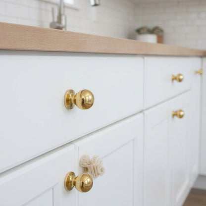 Unlacquered brass ball cabinet knobs on white kitchen drawers and cupboard doors beneath a wood countertop