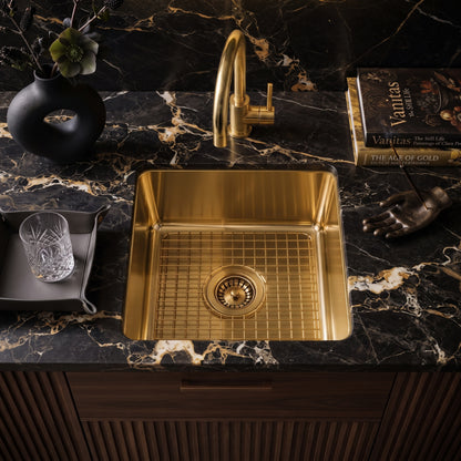 A square brushed brass kitchen sink with a matching undermount faucet, set into a black marble countertop with gold veining. The sink is styled with a glass, a black vase, and a book on a dark wood cabinet.
